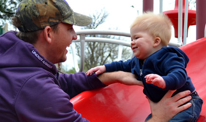 Man in a purple jacket and a baby play at the playground.